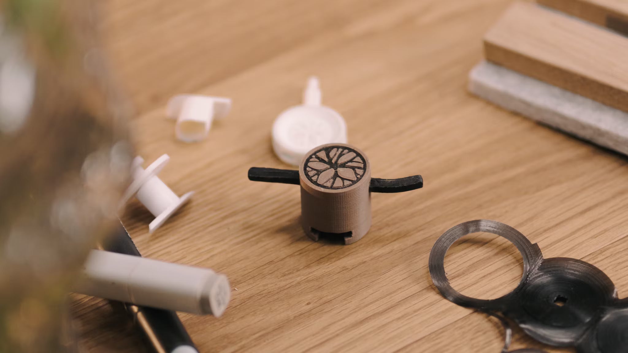 Un petit prototype imprimé en 3D avec des bras noirs est posé sur un bureau en bois à côté de divers composants en plastique, évoquant le développement daccessoires pour panneaux acoustiques.